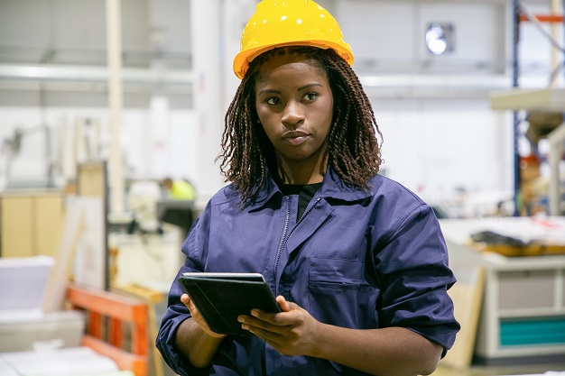 Content female plant worker standing with tablet and looking away. Portrait of pensive African American woman doing her job, thinking and wearing uniform. Manufacture and digital technology concept